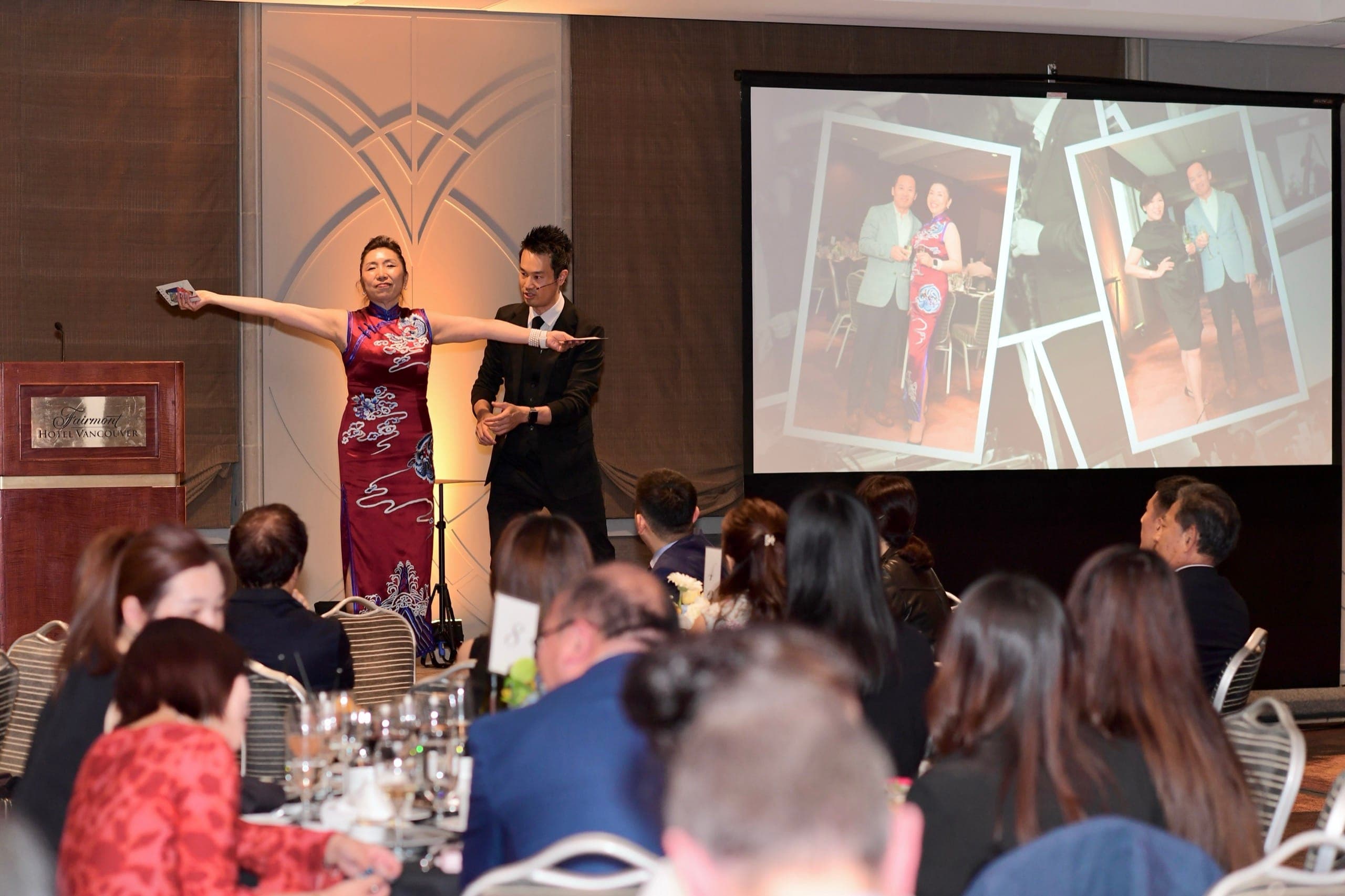 Kel on stage with a volunteer holding up a card while the seated room watches at a Vancouver fundraiser