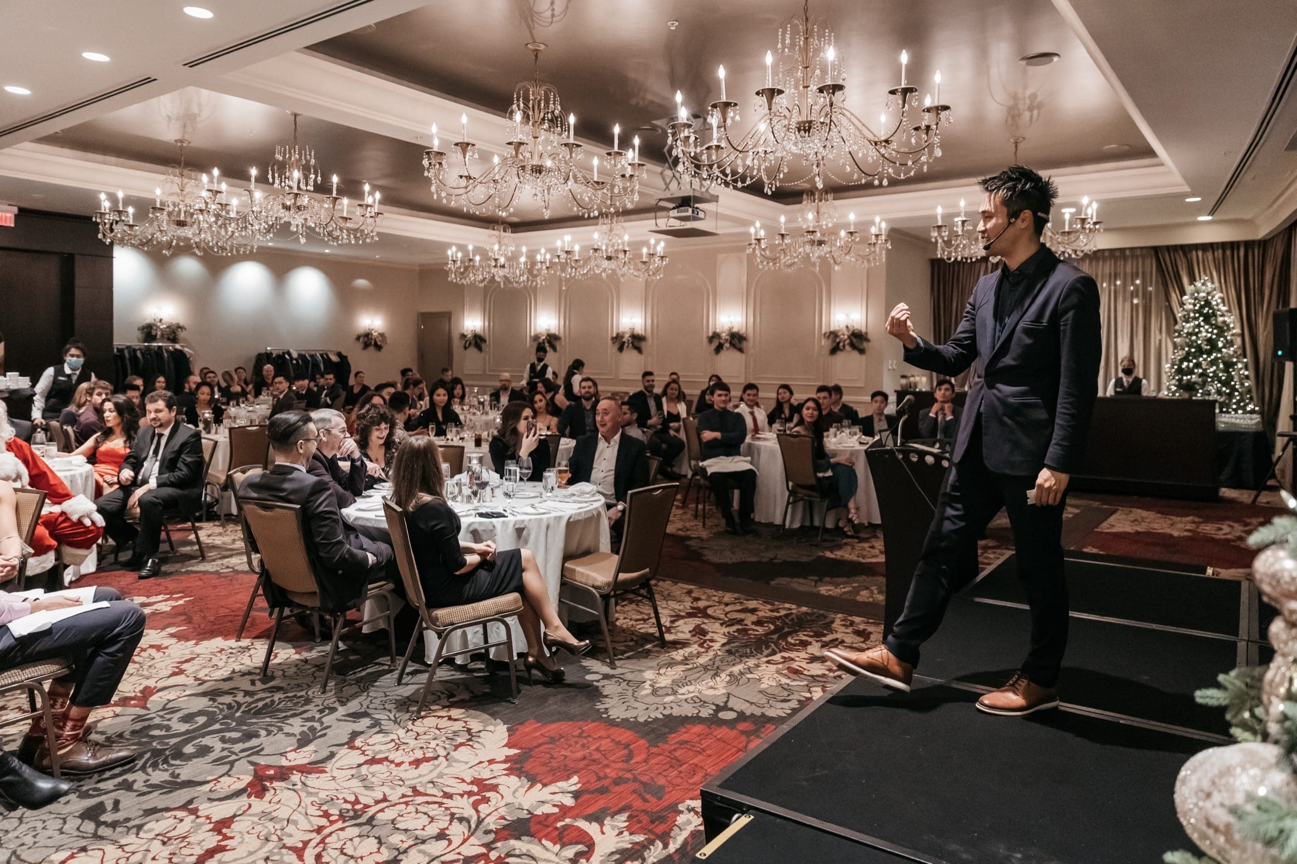 Kel performing a stage show in a full ballroom under chandeliers at a Vancouver corporate holiday party