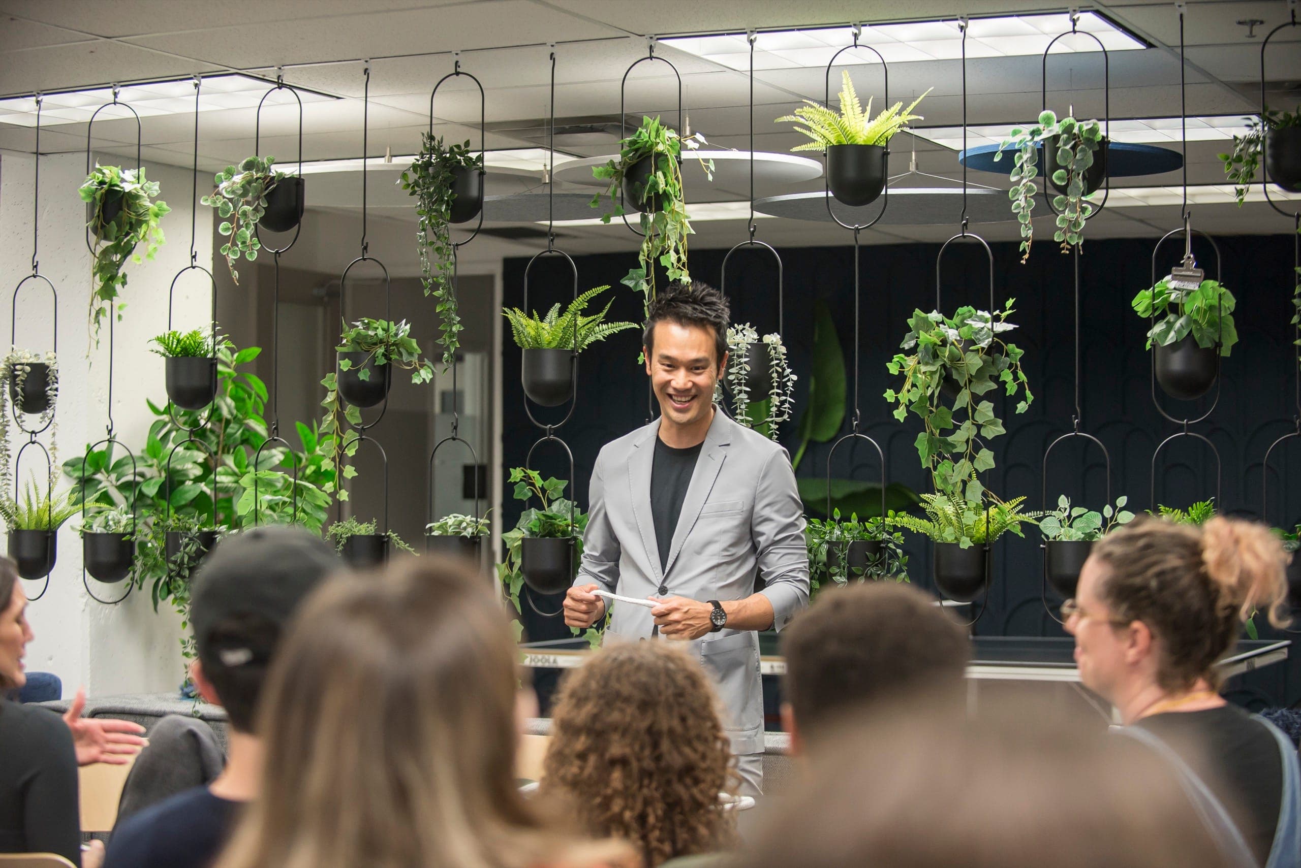 Kel laughing mid-routine in front of a wall of hanging planters at a corporate event