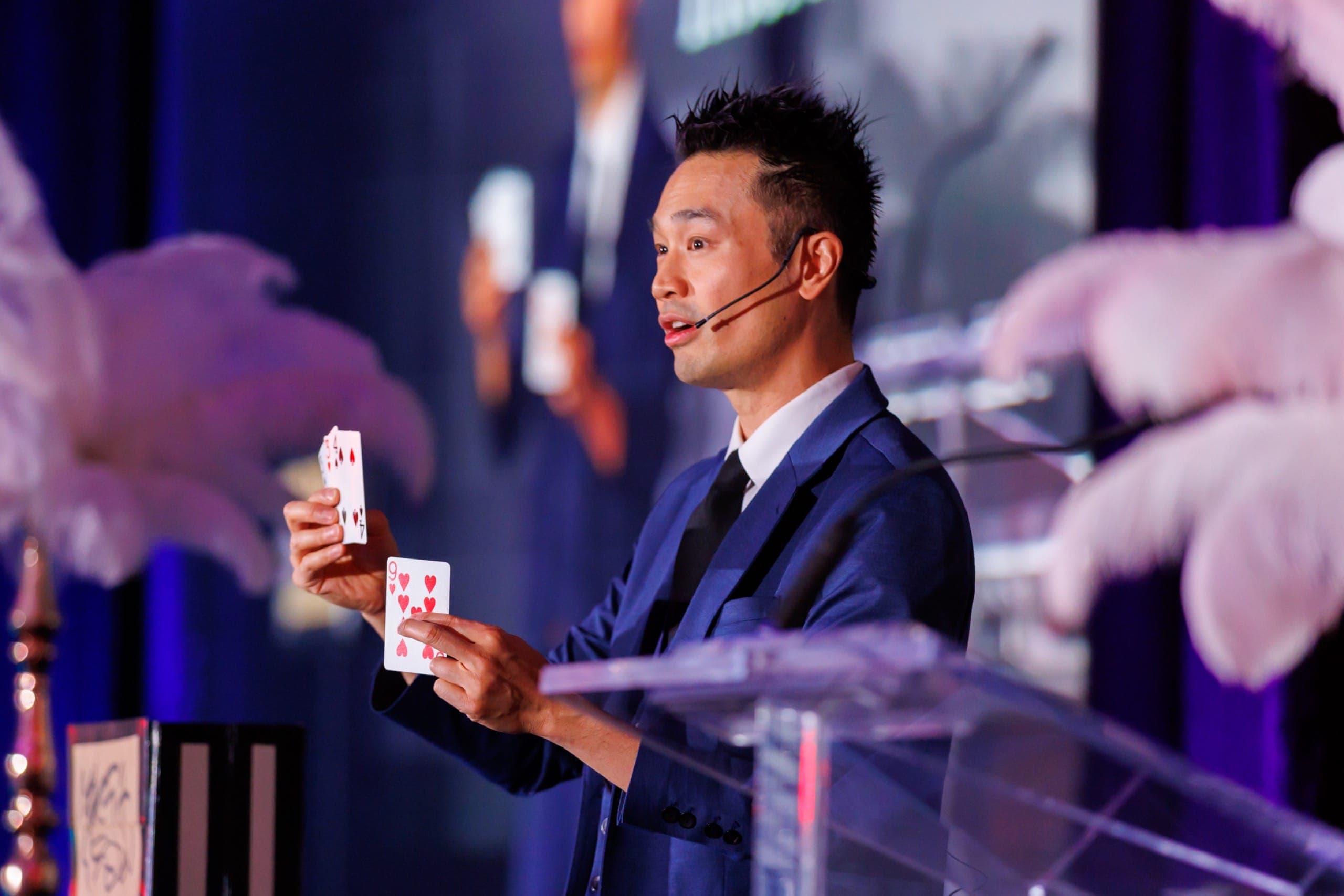 Kel on stage under blue lighting holding up two cards during the after-dinner show at a Hyatt Regency gala