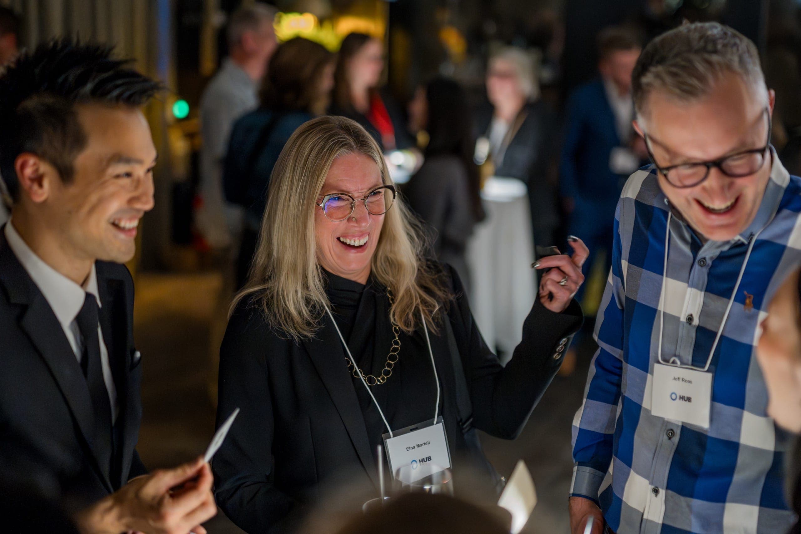 Kel laughing with two guests during close-up magic at a Vancouver firm's holiday reception, the room busy behind them