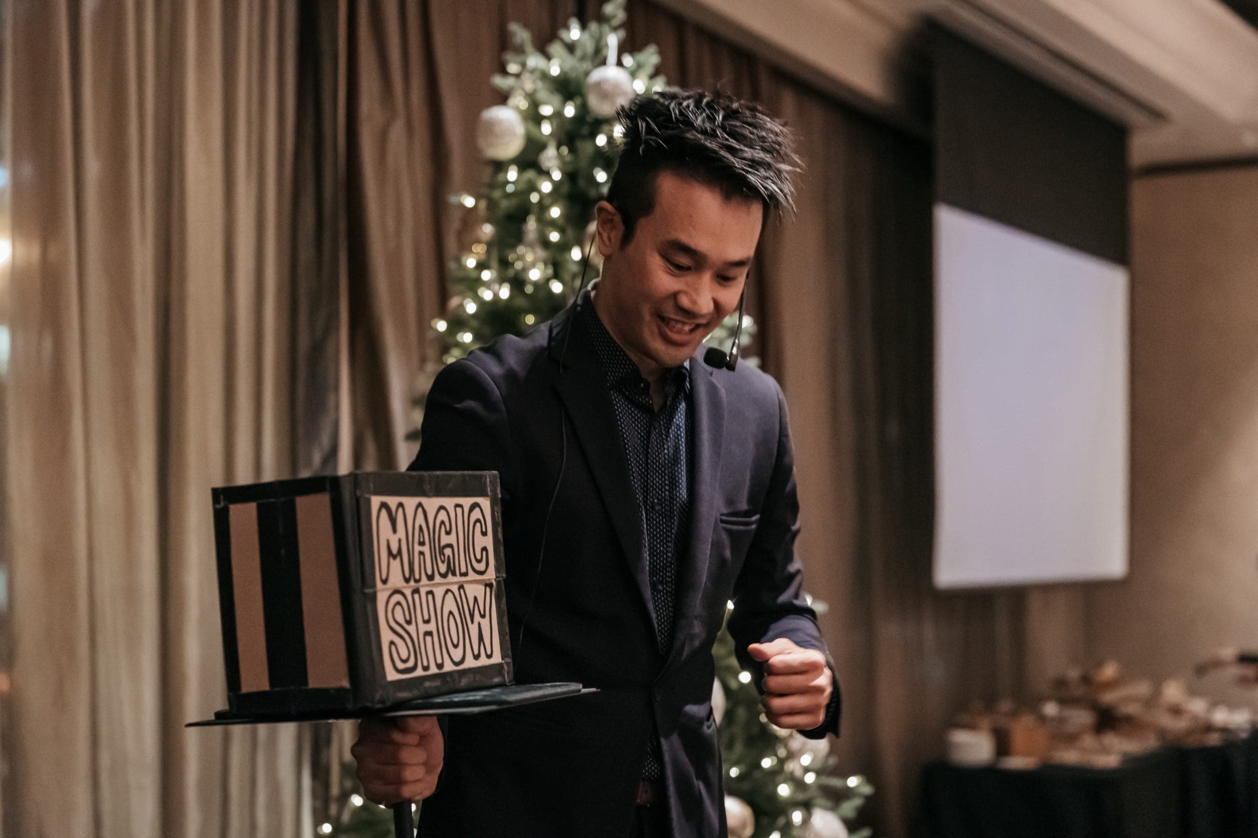 Kel smiling and holding his hand-lettered magic show box beside a Christmas tree at a Vancouver event