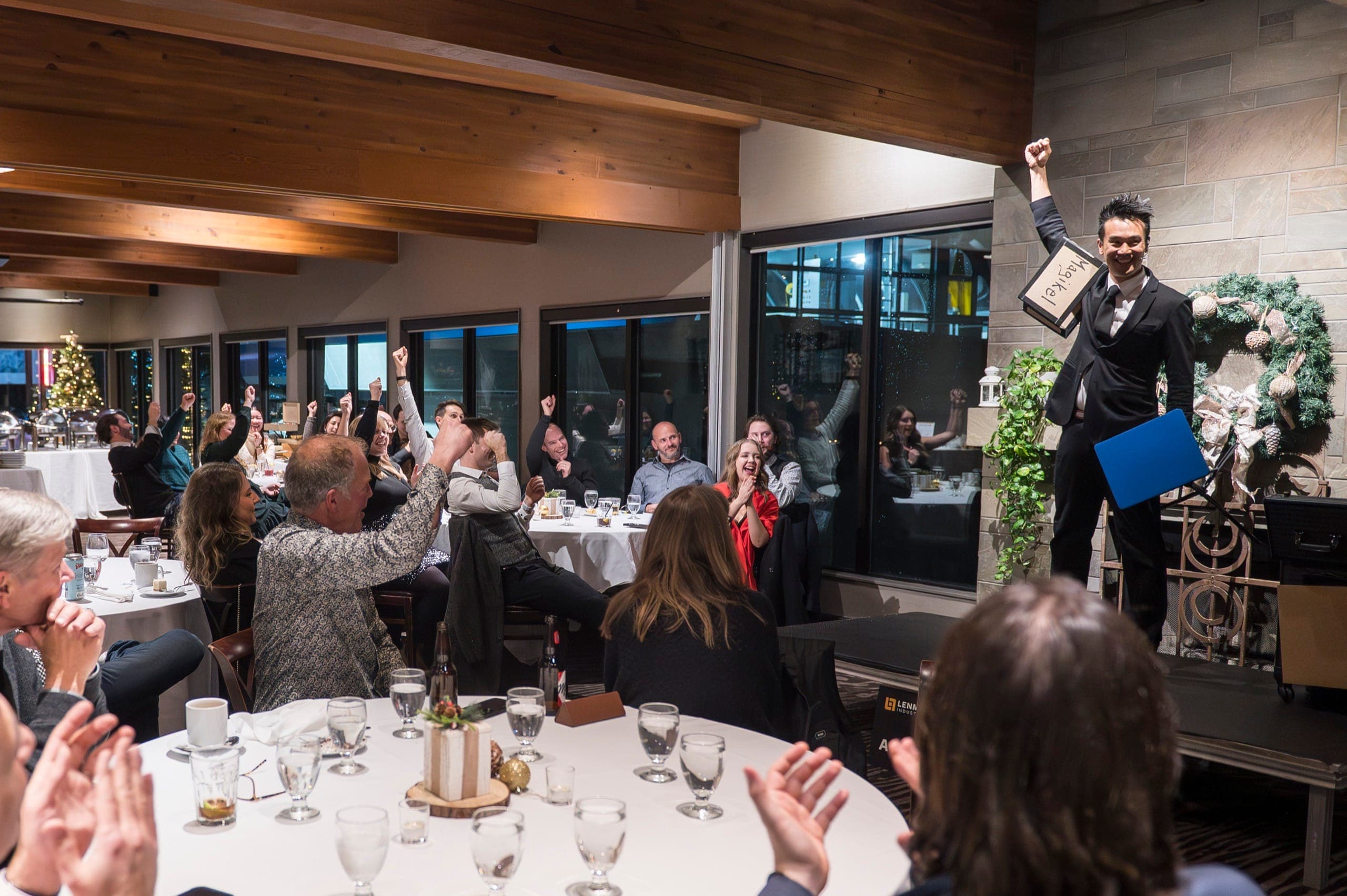 Kel raising a sign on stage while the room cheers with arms up at a Vancouver corporate event