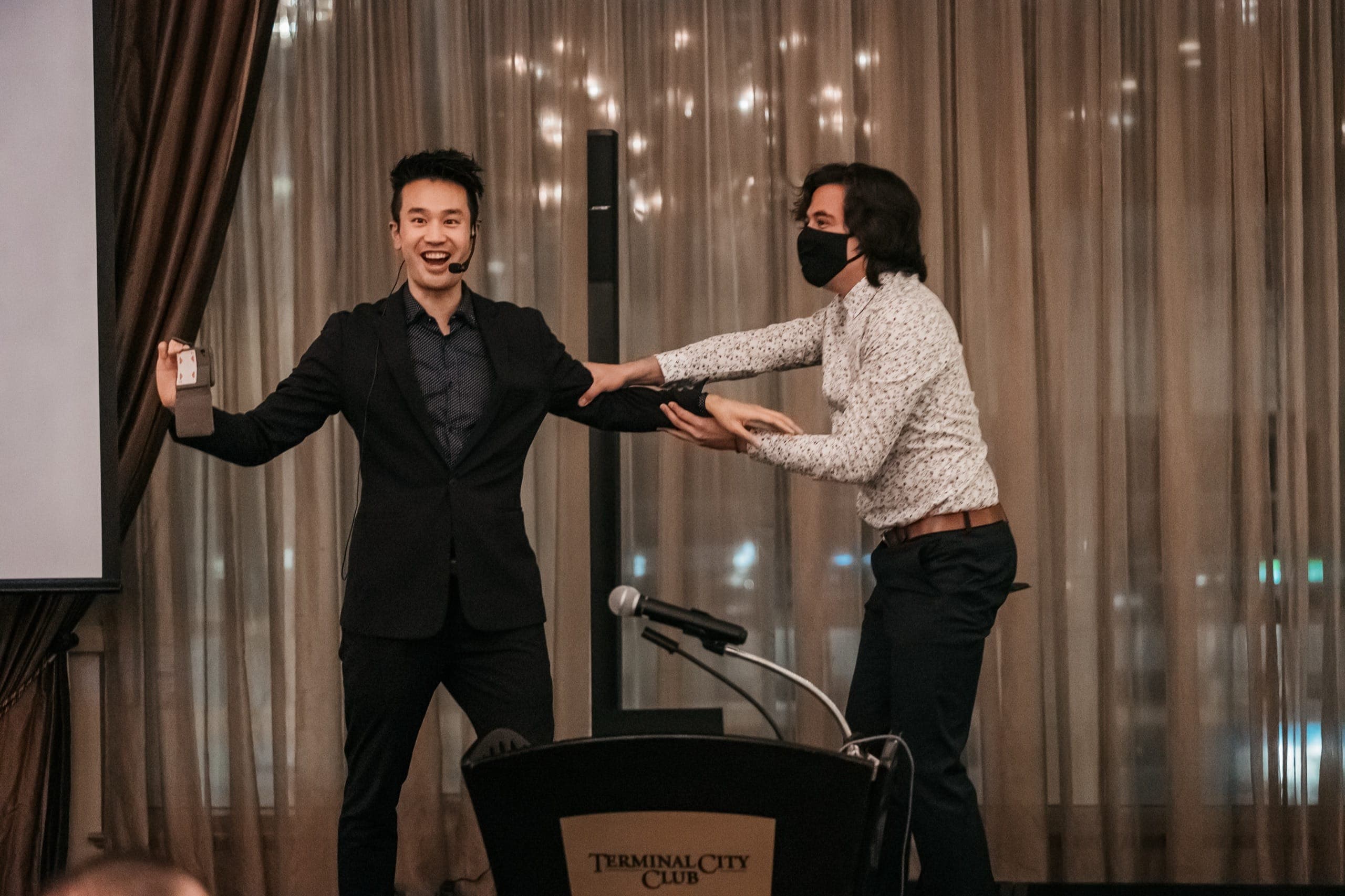 Kel hosting from the podium at the Terminal City Club in Vancouver, sharing a laugh with a guest on stage