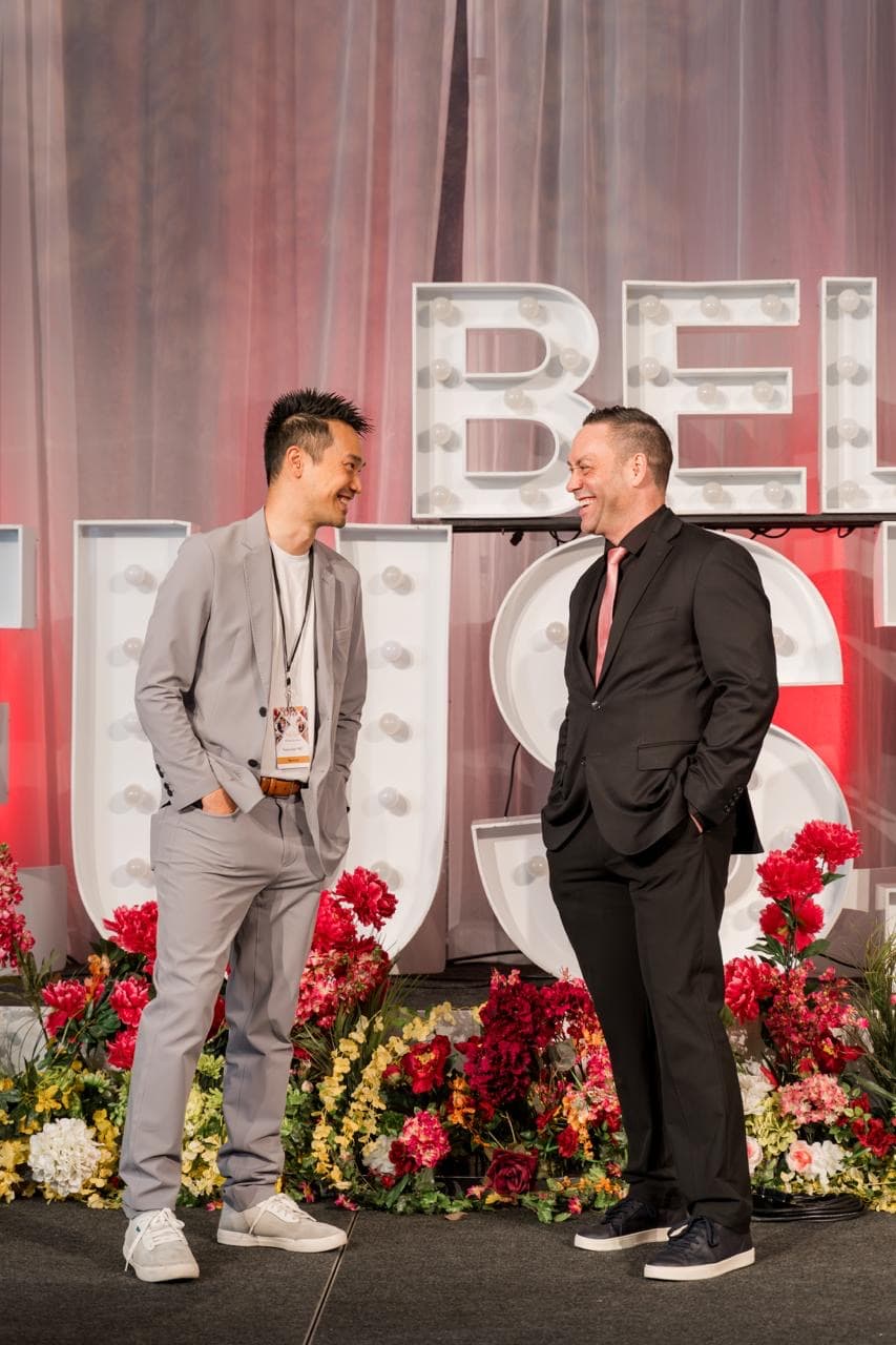 Kel and Mike Oulton laughing together on stage in front of lit marquee letters and a bank of red flowers