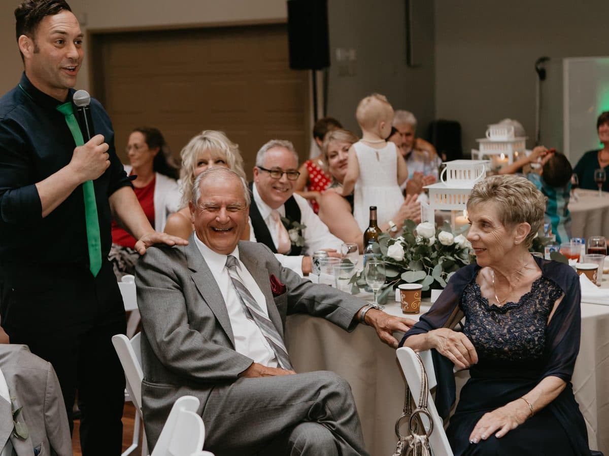 Mike Oulton hosting with a microphone beside a table of laughing guests at a Vancouver event