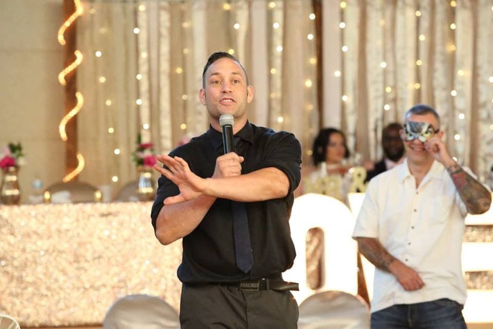 Mike Oulton hosting with a handheld microphone mid-gesture in front of a lit reception backdrop