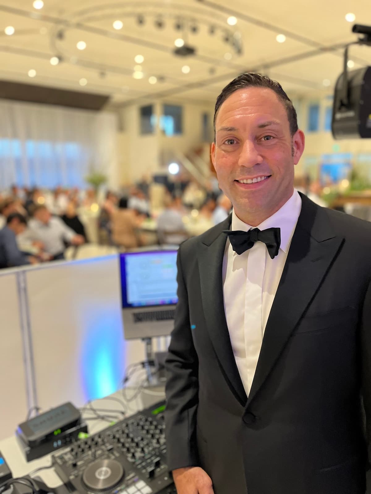 Mike Oulton in a tuxedo at the DJ booth with the ballroom and guests behind him at a Vancouver black-tie event