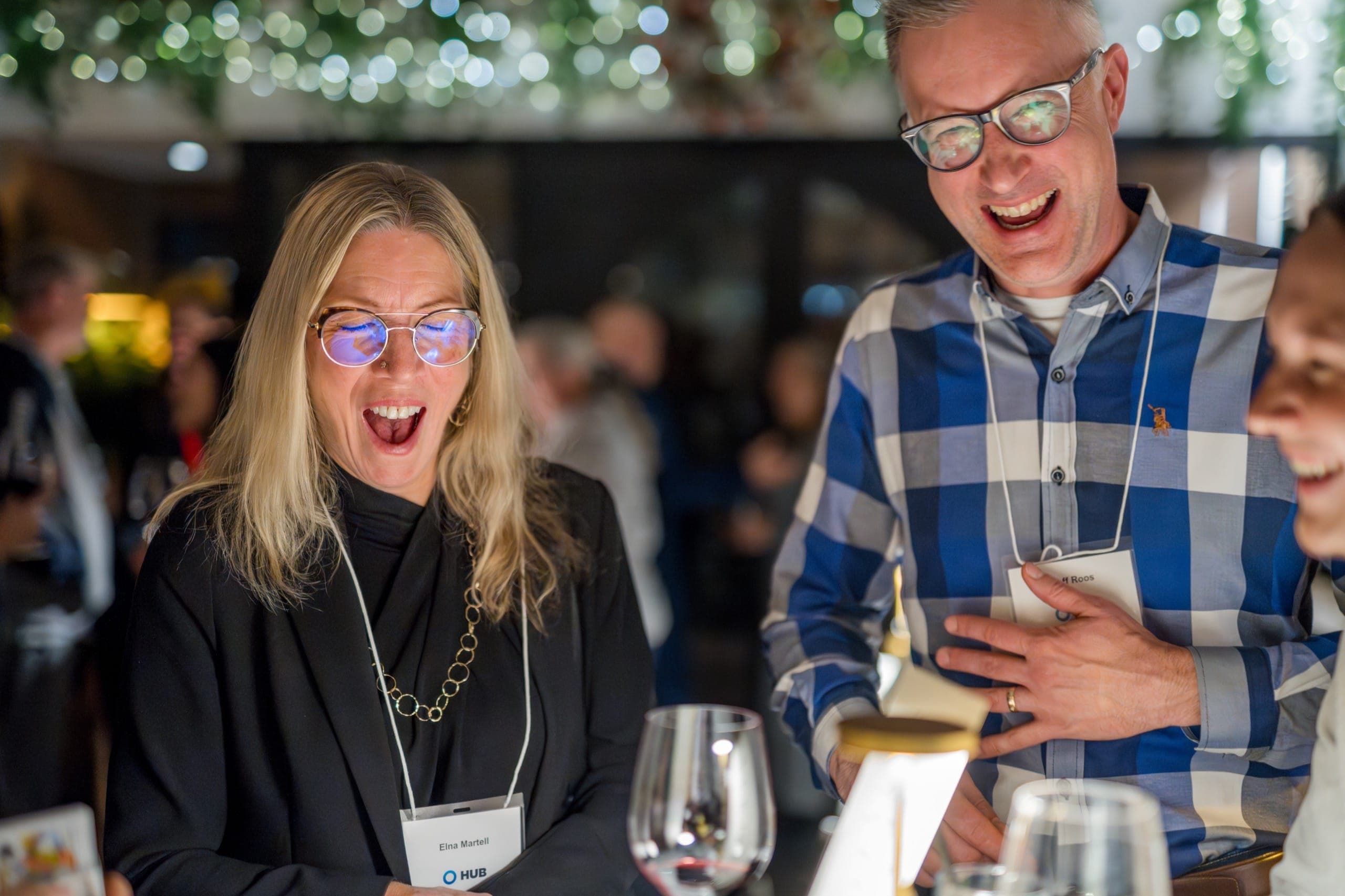 One guest gasping open-mouthed while another doubles over laughing during close-up magic at a Vancouver corporate holiday party