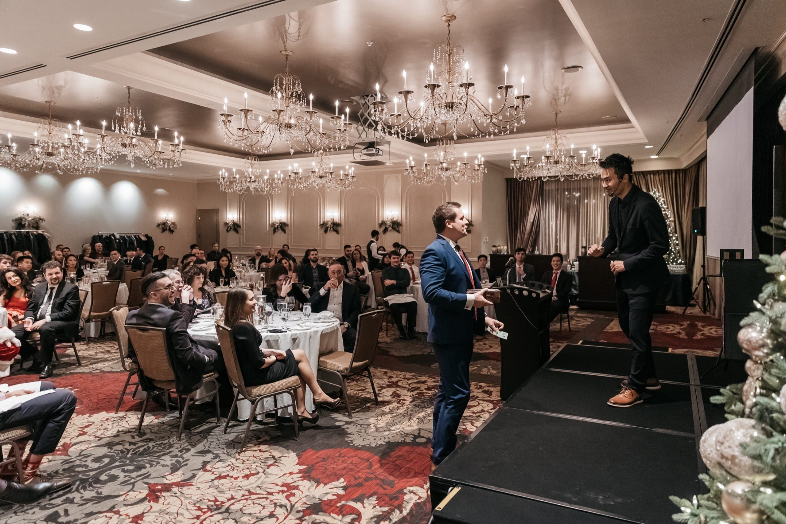 Kel performing with a volunteer in a packed ballroom under crystal chandeliers at a Vancouver corporate gala