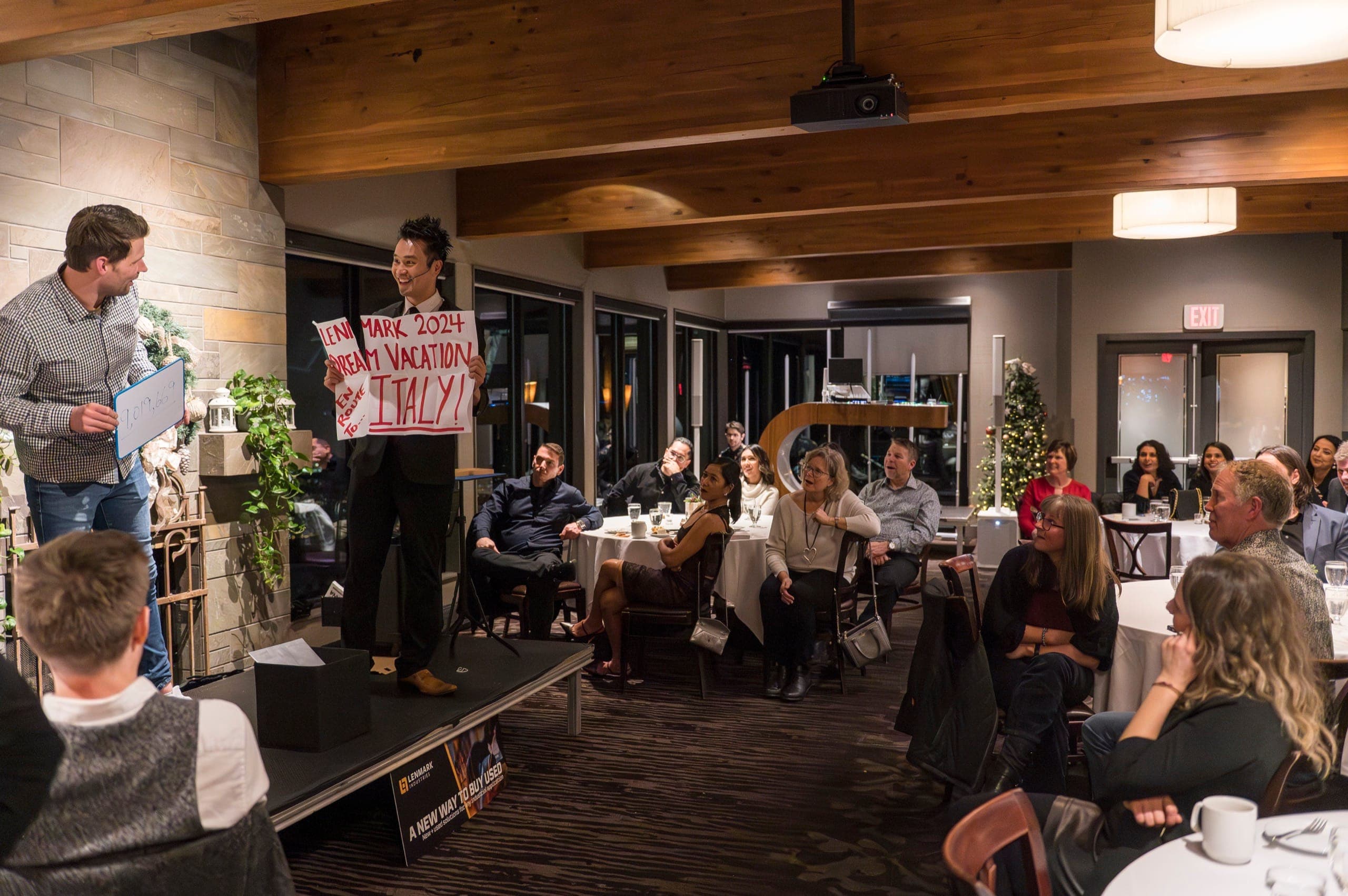 Kel revealing a written prediction on stage while the whole room watches at a Vancouver corporate dinner