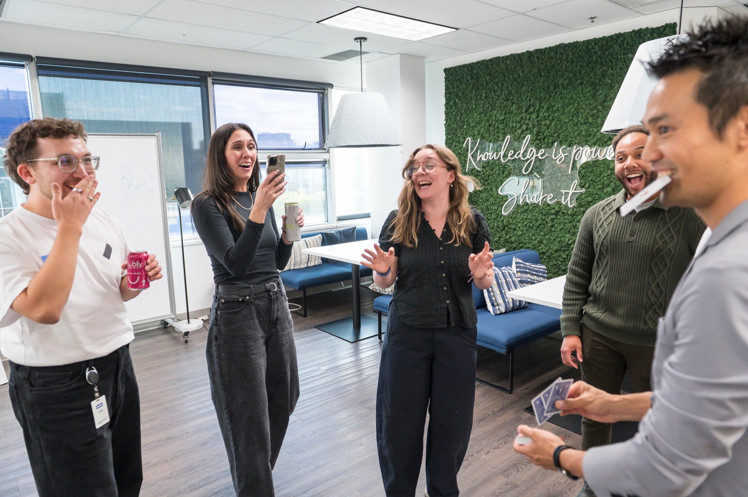 Three colleagues reacting at once, one filming on her phone, during close-up magic at a Vancouver tech event