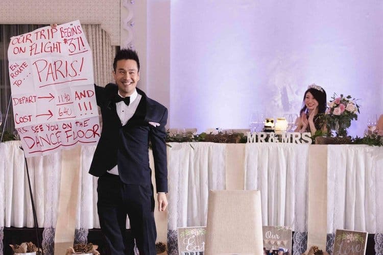 Kel holding up a handwritten honeymoon destination prediction beside the newlyweds' sweetheart table