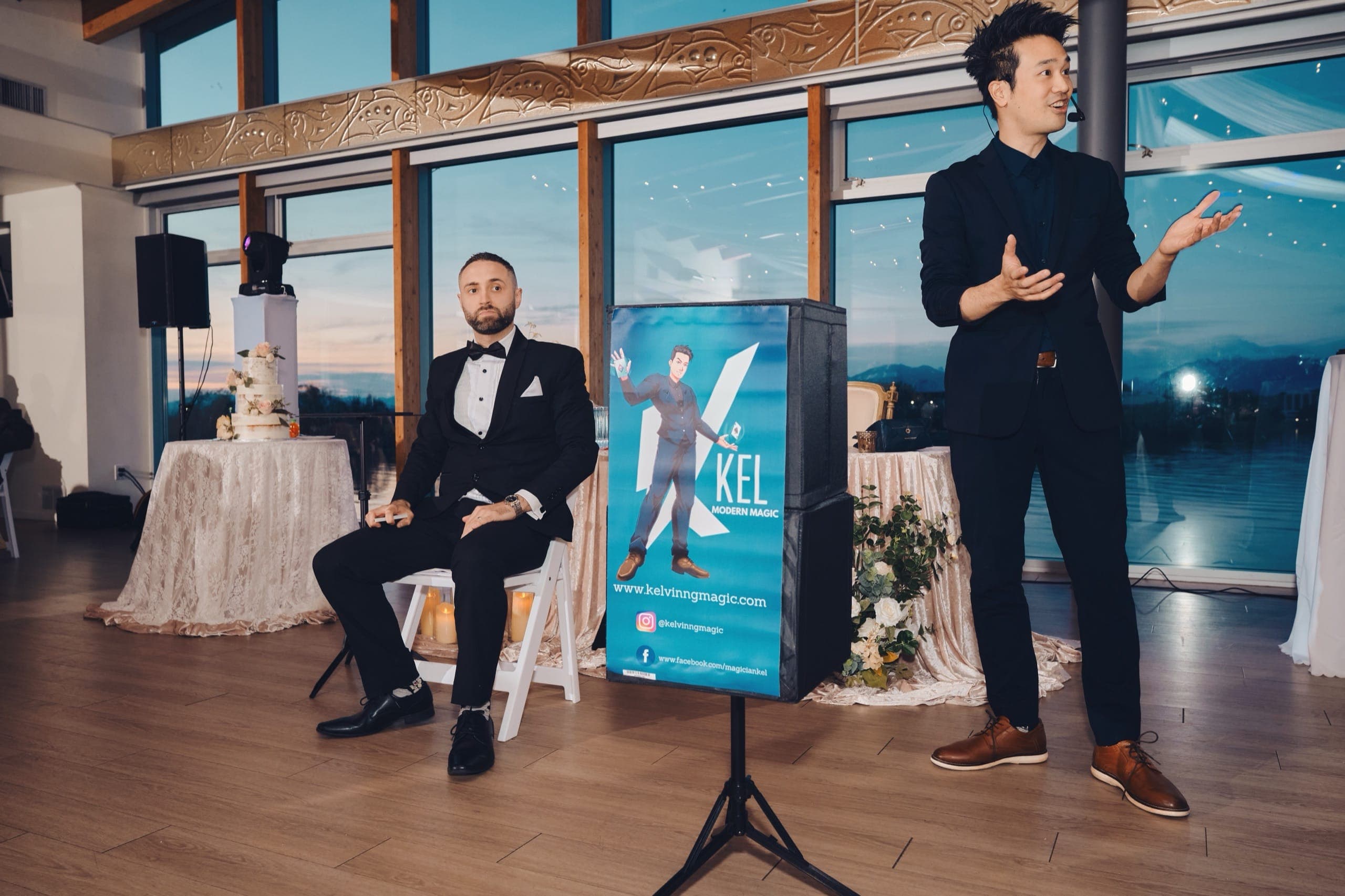 Kel hosting a wedding reception as MC beside a branded Kel Modern Magic sign, sunset venue view