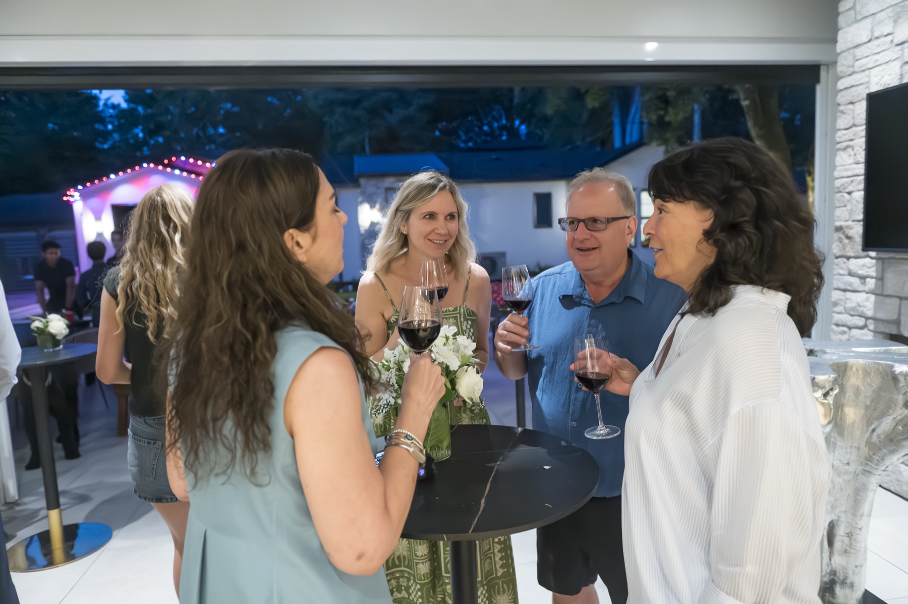Guests talking over drinks beside open glass walls as a Vancouver corporate summer party moves from daylight into evening