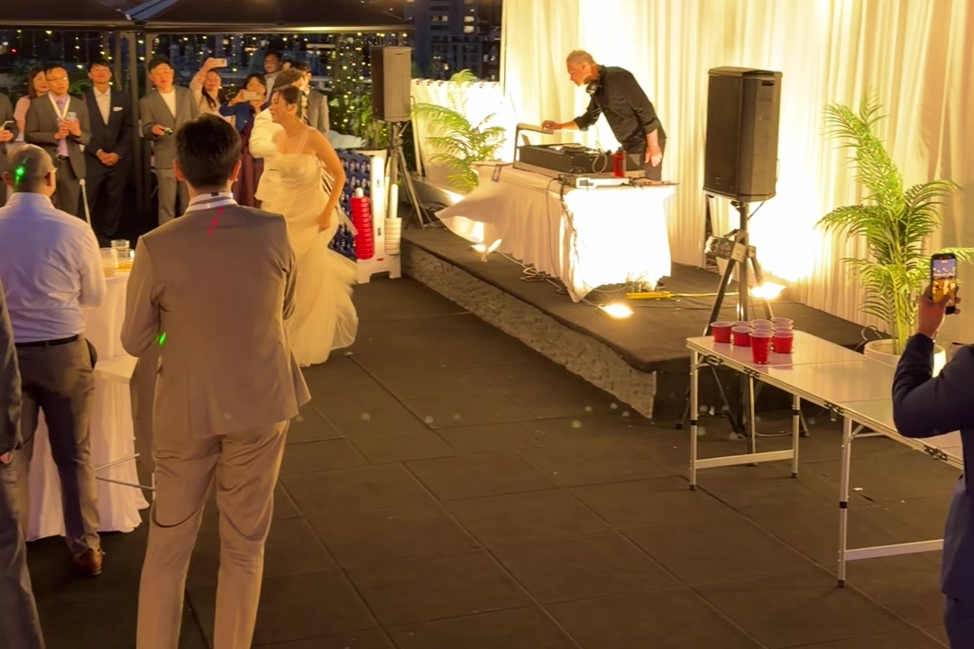 A DJ working the decks on a draped rooftop riser at dusk, the bride nearby and the Vancouver skyline behind