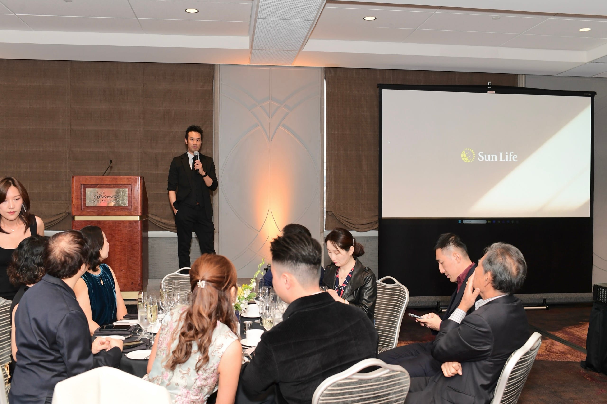 Kel addressing a seated ballroom with a handheld microphone during a Vancouver corporate event